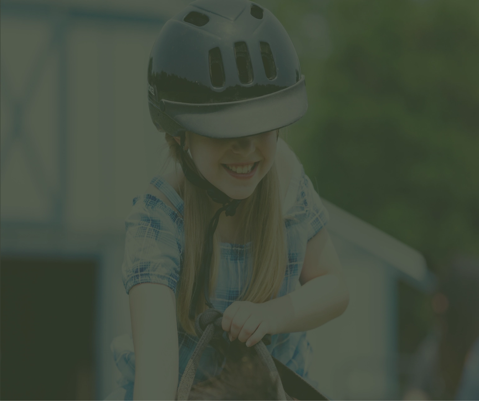A smiling young girl in a riding helmet and blue plaid shirt enjoys a horseback ride near the barn.