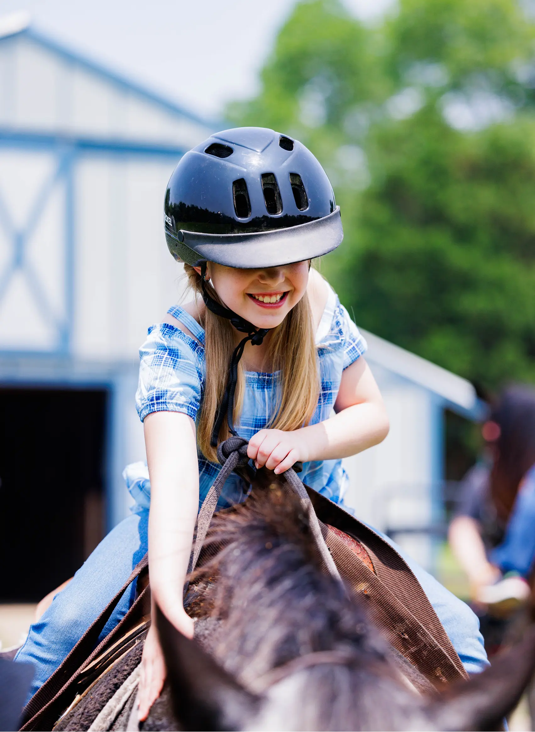 A smiling girl in a riding helmet and plaid shirt rides a brown horse near the ranch barn.