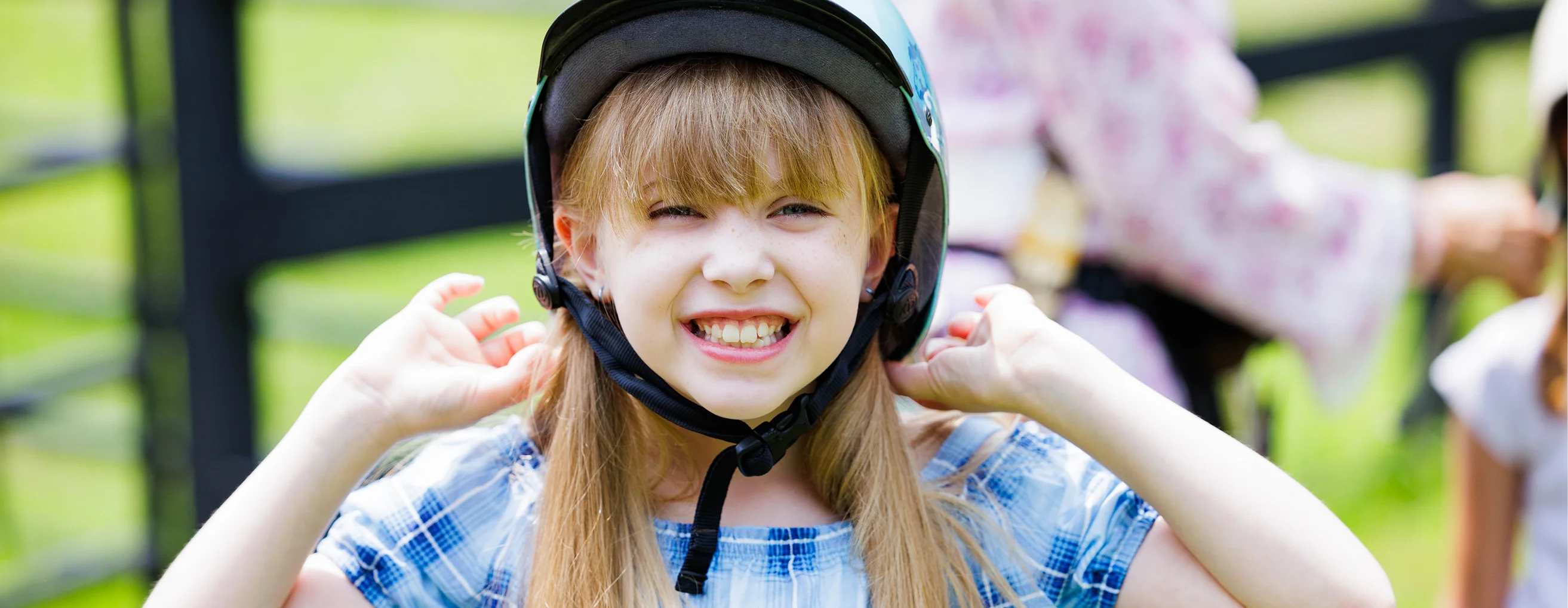 A smiling young girl with blonde bangs adjusts a teal riding helmet outdoors near a pasture fence.
