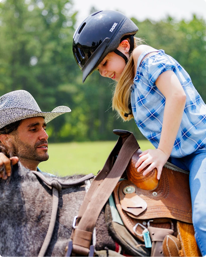 A man in a cowboy hat helps a smiling girl in a riding helmet sitting on a dark horse in a sunny field.