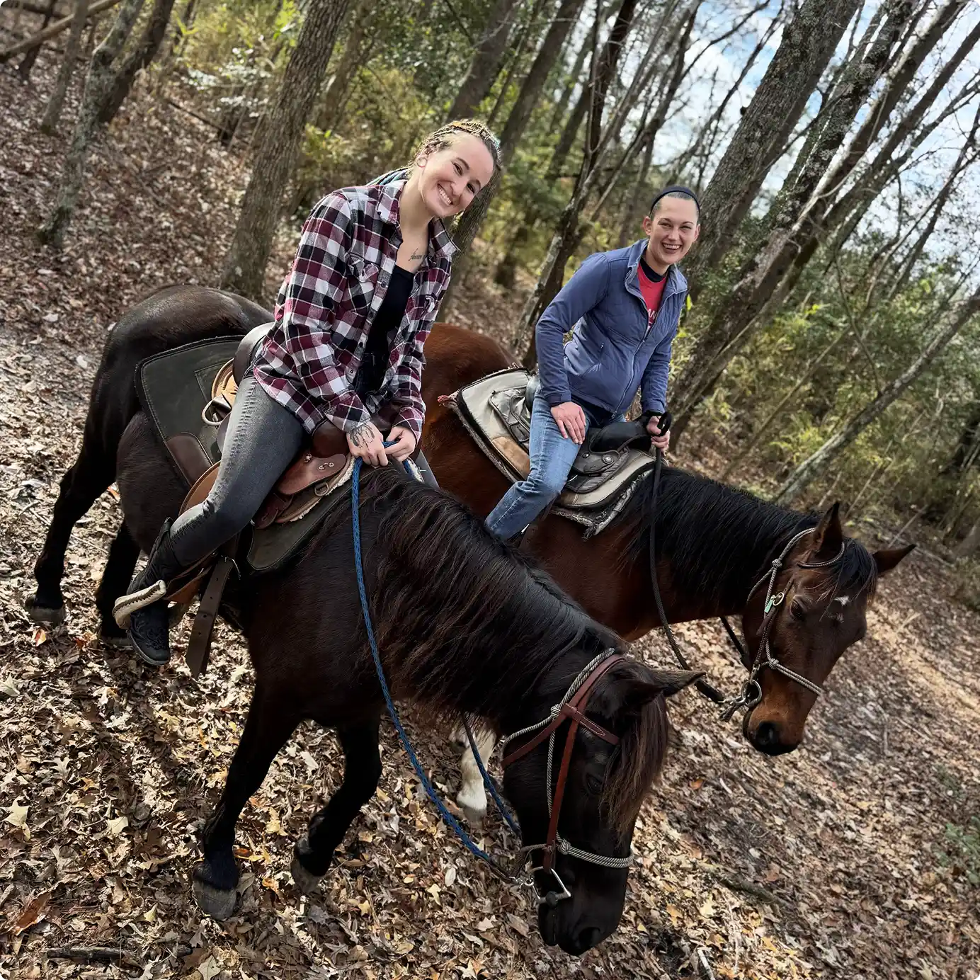 Two smiling riders on horseback follow a leaf-covered trail through the woods.