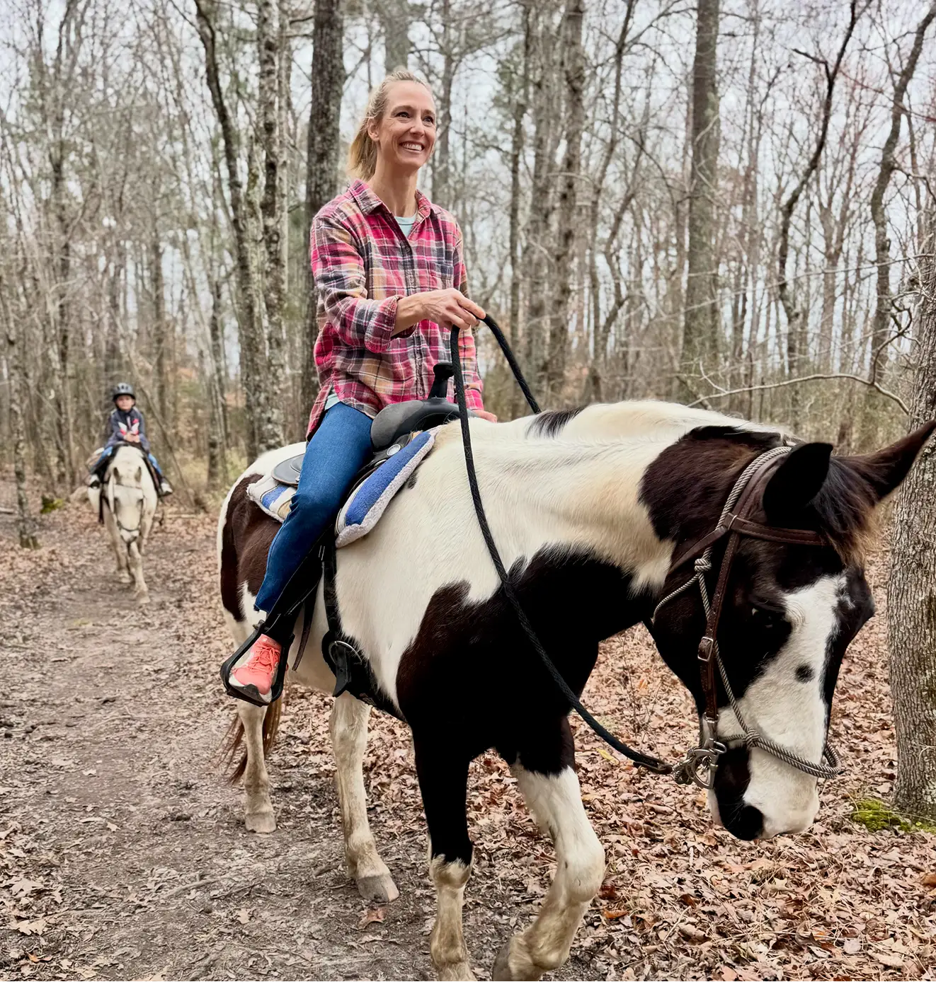 A smiling woman on a pinto horse leads a child on a light horse along a wooded trail.