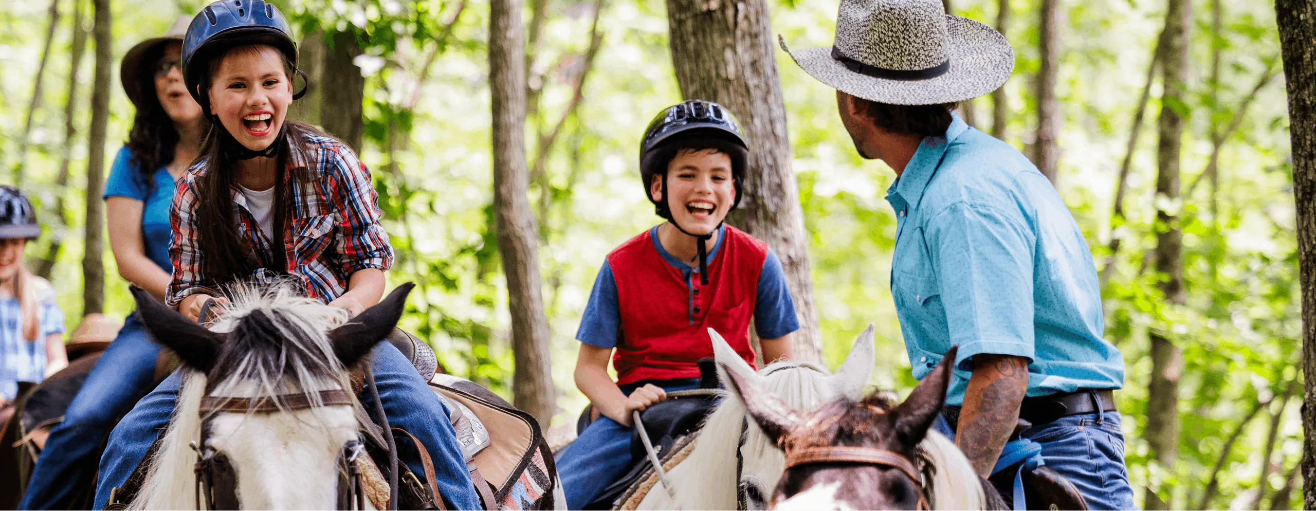 Children in riding helmets and a guide in a cowboy hat laugh while riding horses through a wooded trail.