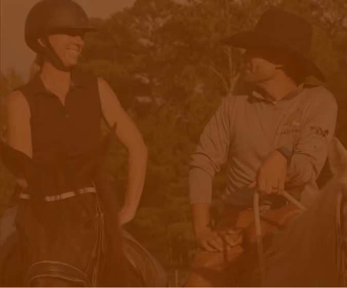 A woman in a riding helmet and a man in a cowboy hat smile at each other on horseback in the woods.