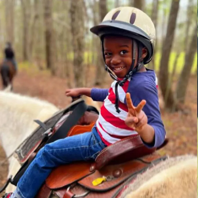 A smiling child in a riding helmet makes a peace sign while riding a light-colored horse on a wooded trail.