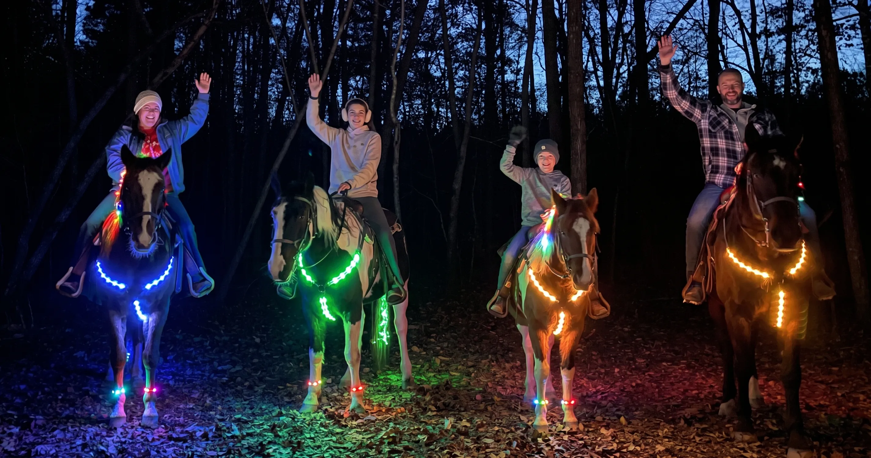 A family of four waves while riding horses at night, each horse outlined in blue, green, or orange LED lights.