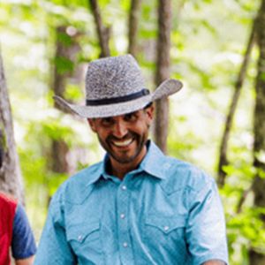 A smiling Kay's Acres trainer in a cowboy hat and light-blue western shirt stands among trees.
