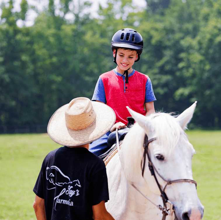 A smiling child in a riding helmet sits on a white horse while a Kay's Acres guide in a straw hat stands beside them.
