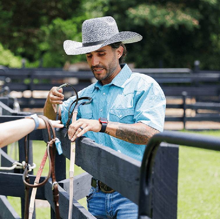 A bearded trainer in a cowboy hat and western shirt adjusts leather tack over a black paddock fence.