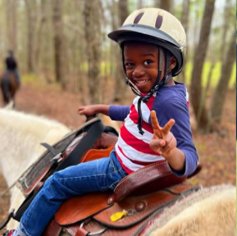 A smiling child in a riding helmet makes a peace sign while riding a light-colored horse on a wooded trail.