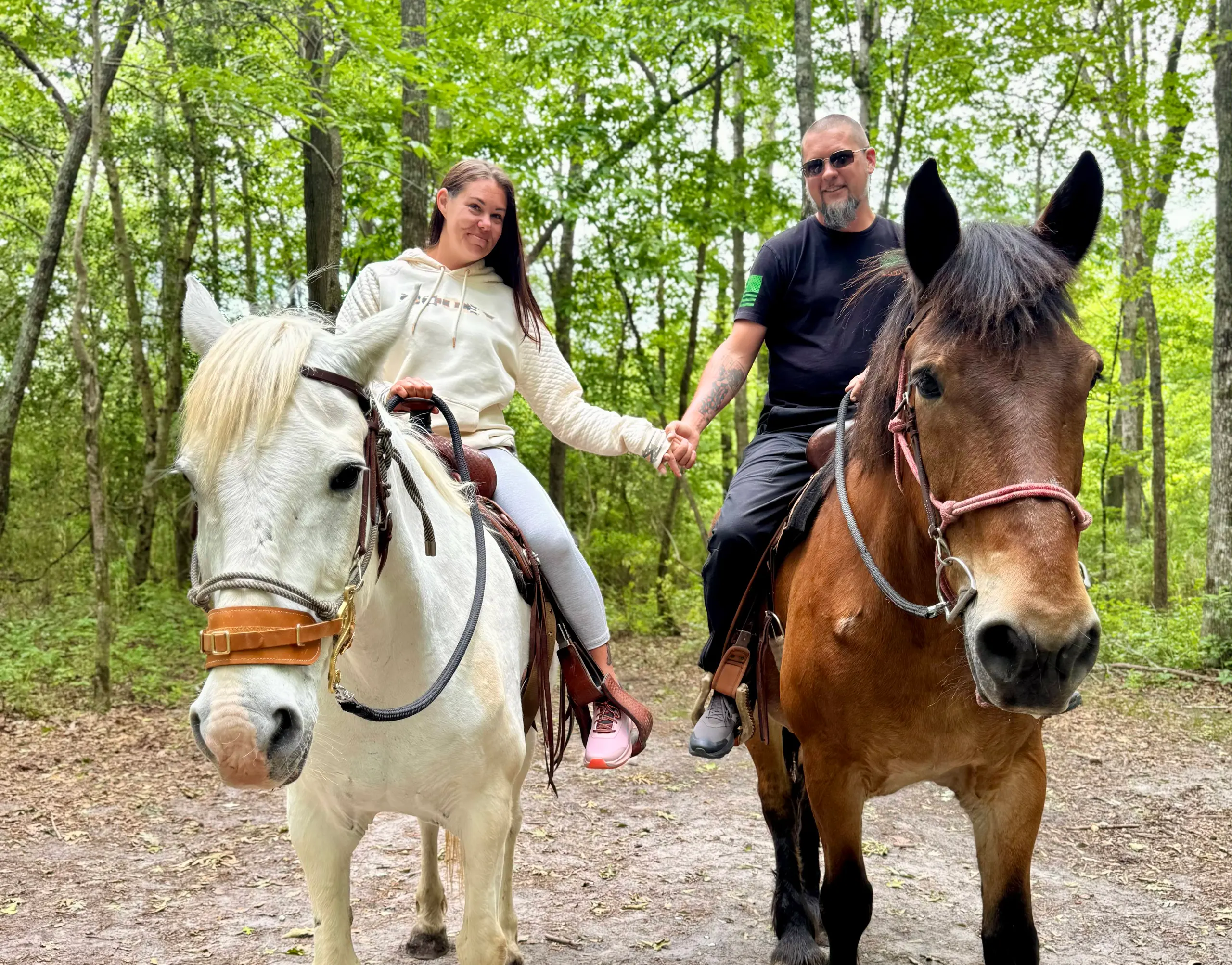 A couple holds hands while riding side by side on a wooded trail, one on a white horse and one on a brown horse.