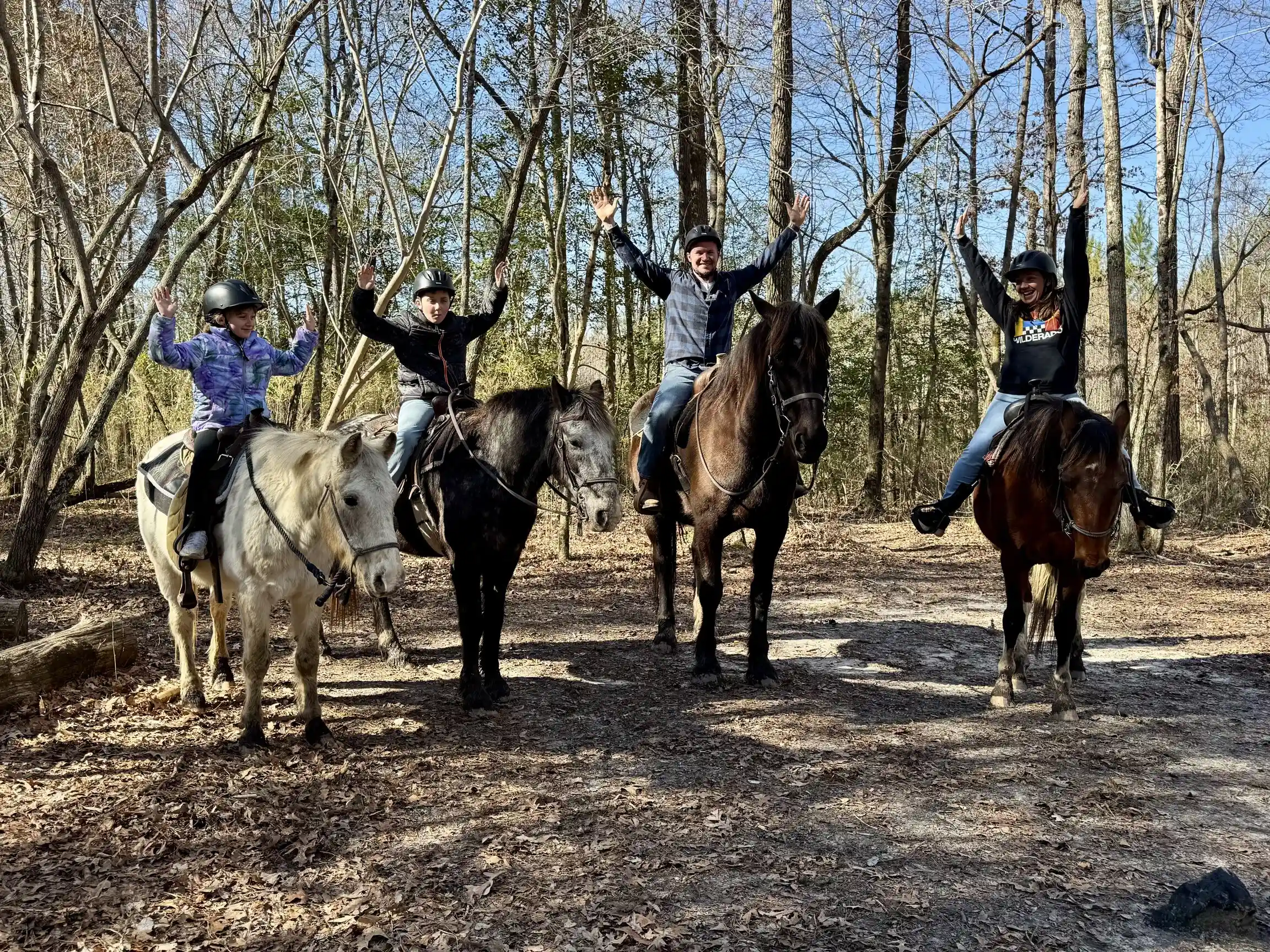 A family of four raises their arms while riding horses on a leaf-covered woodland trail.