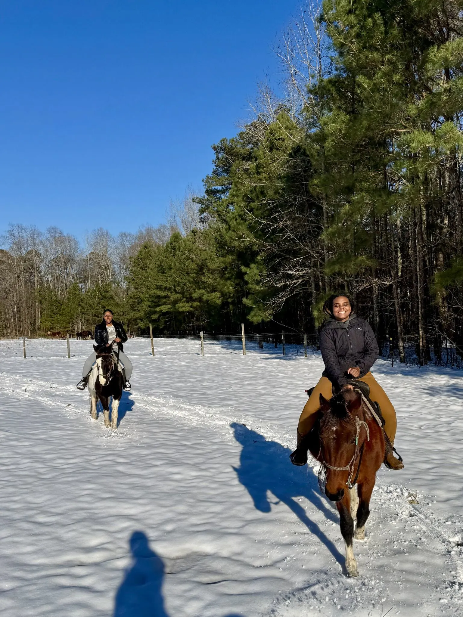 A smiling woman rides a brown horse toward the camera across a snowy field, with a second rider behind.