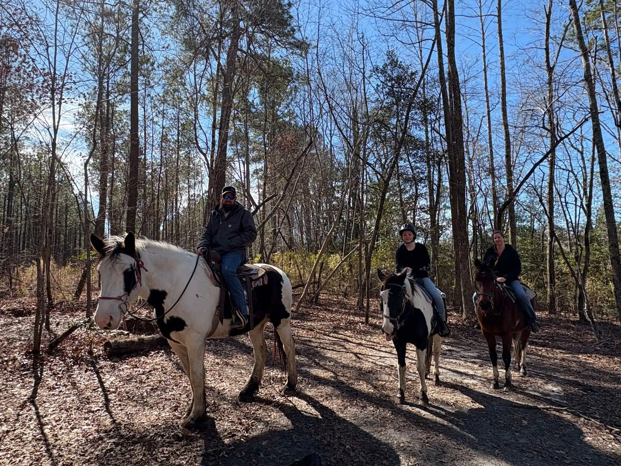 Three people ride horses along a leaf-strewn forest trail under a bright blue sky.