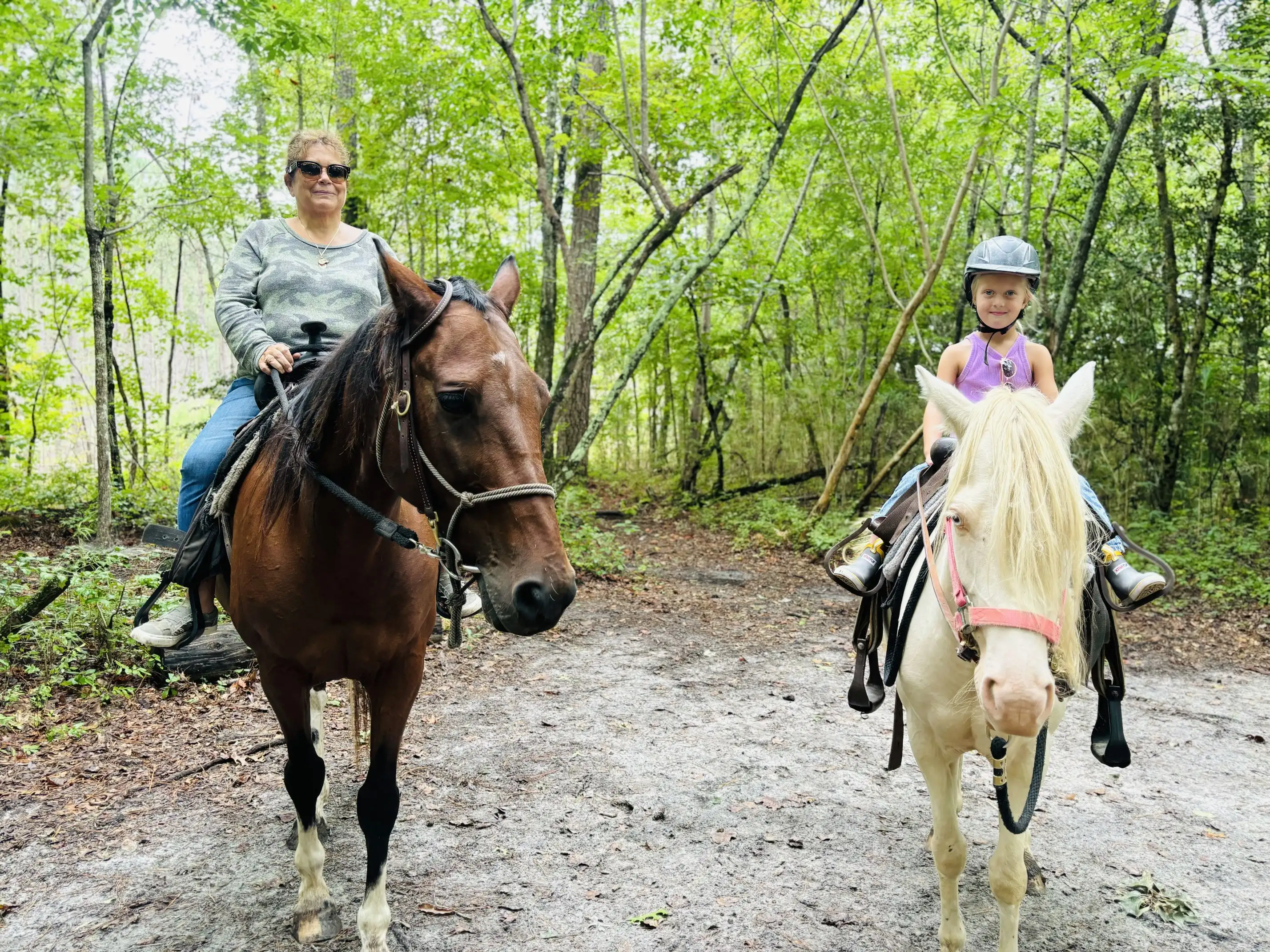 A woman on a brown horse rides beside a smiling girl in a helmet on a white horse along a wooded trail.