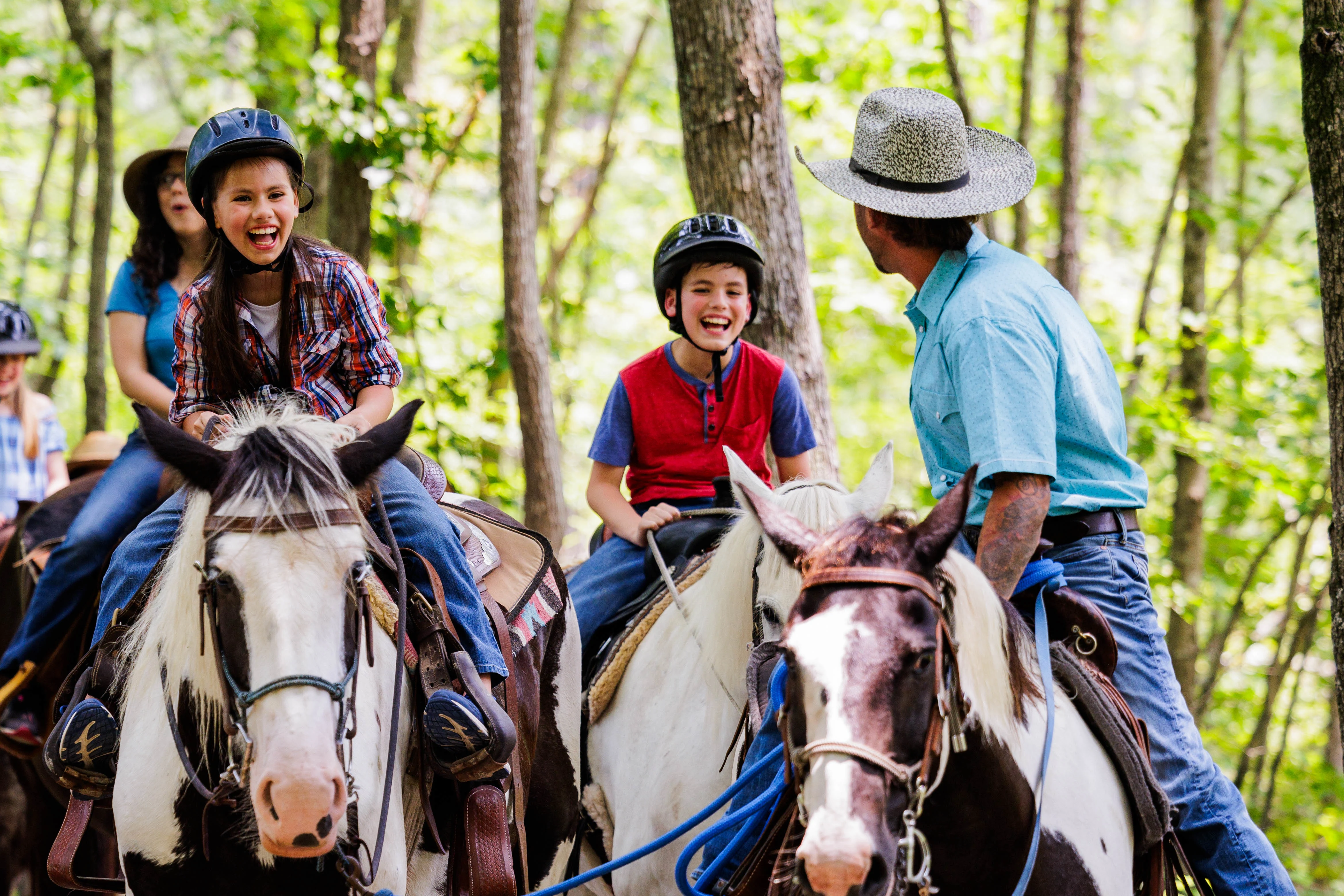 Laughing children in riding helmets ride paint and white horses on a sunlit forest trail with a guide in a cowboy hat.