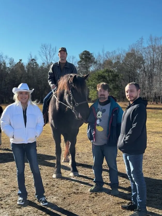 Four people pose with a large dark horse in a sunny open field under a clear blue sky.