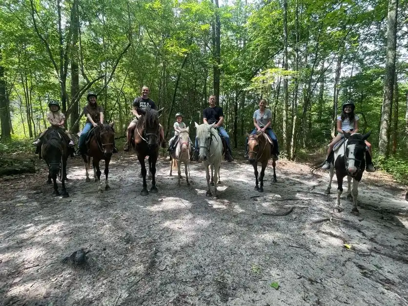 Seven riders, including children in helmets, pose on horseback along a sandy trail in a green forest.