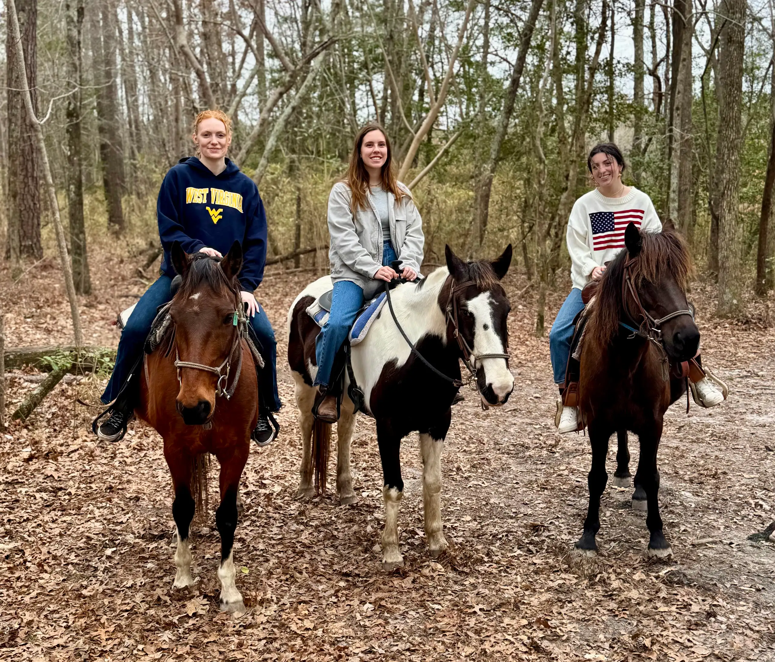 Three women pose on horseback among bare trees and fallen leaves on a woodland trail.