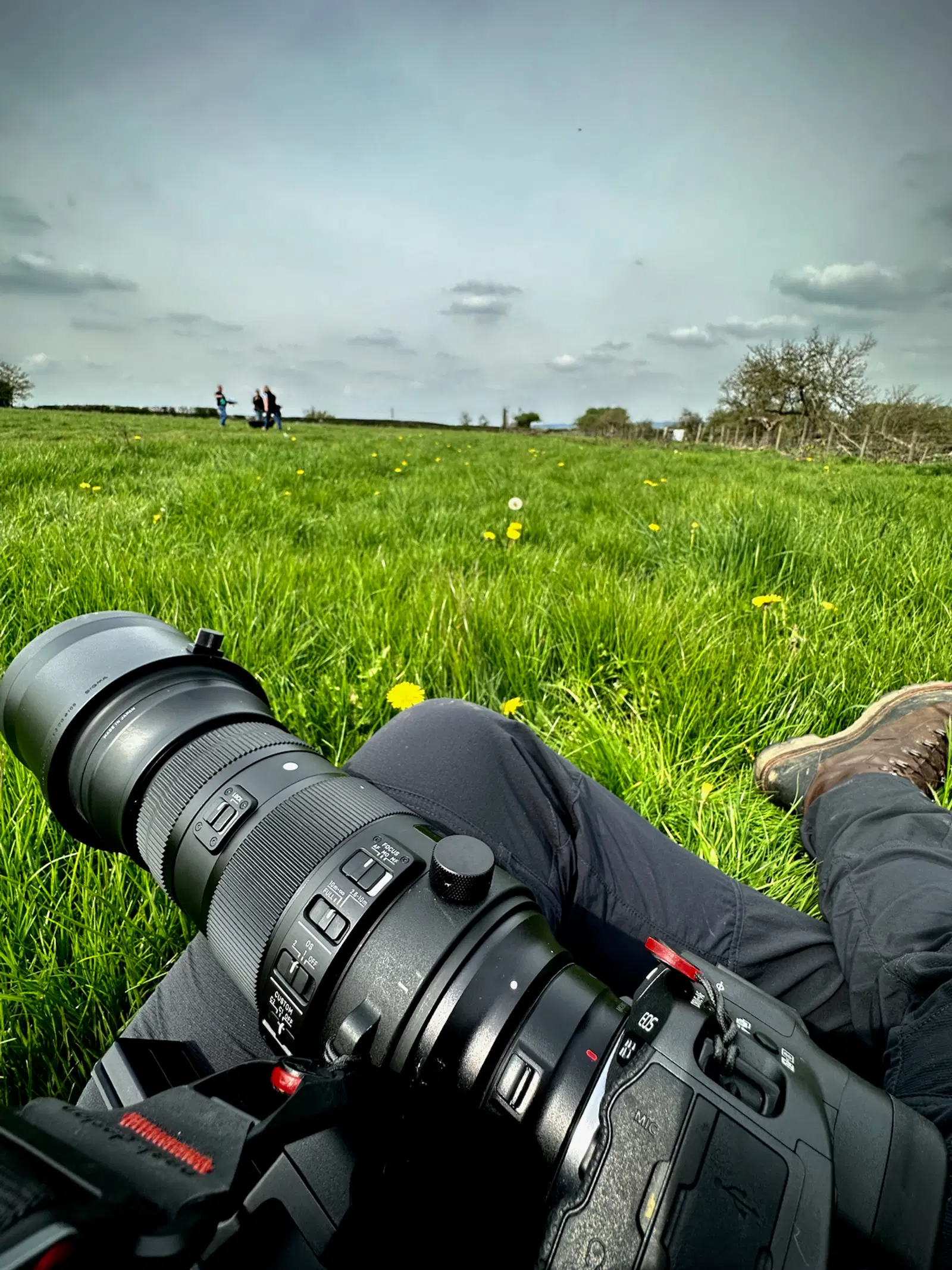 Close-up of a professional DSLR camera with a telephoto lens resting on a photographer’s lap in a grassy field