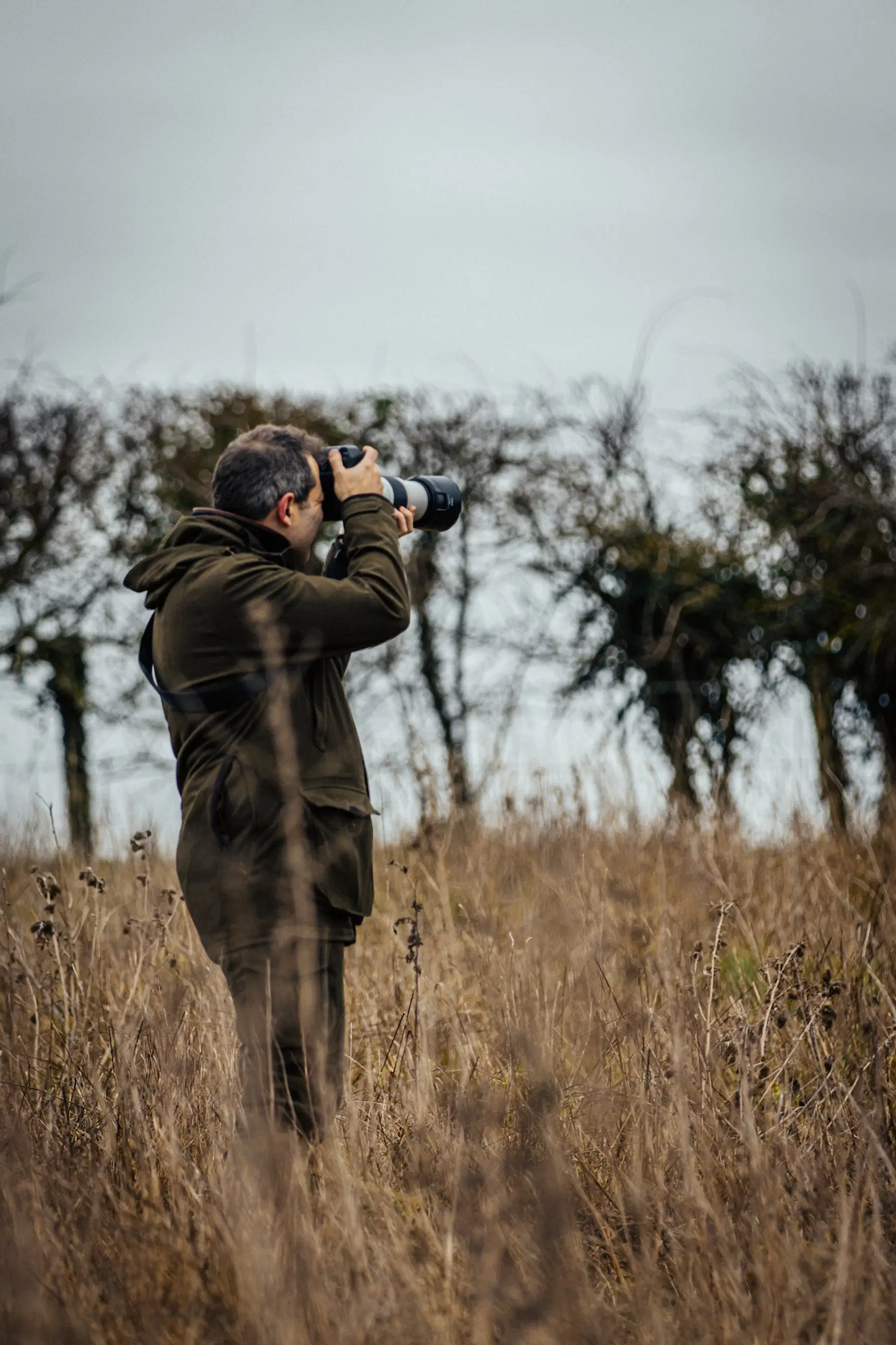 A photographer dressed in olive outdoor gear stands in a field aiming a telephoto lens