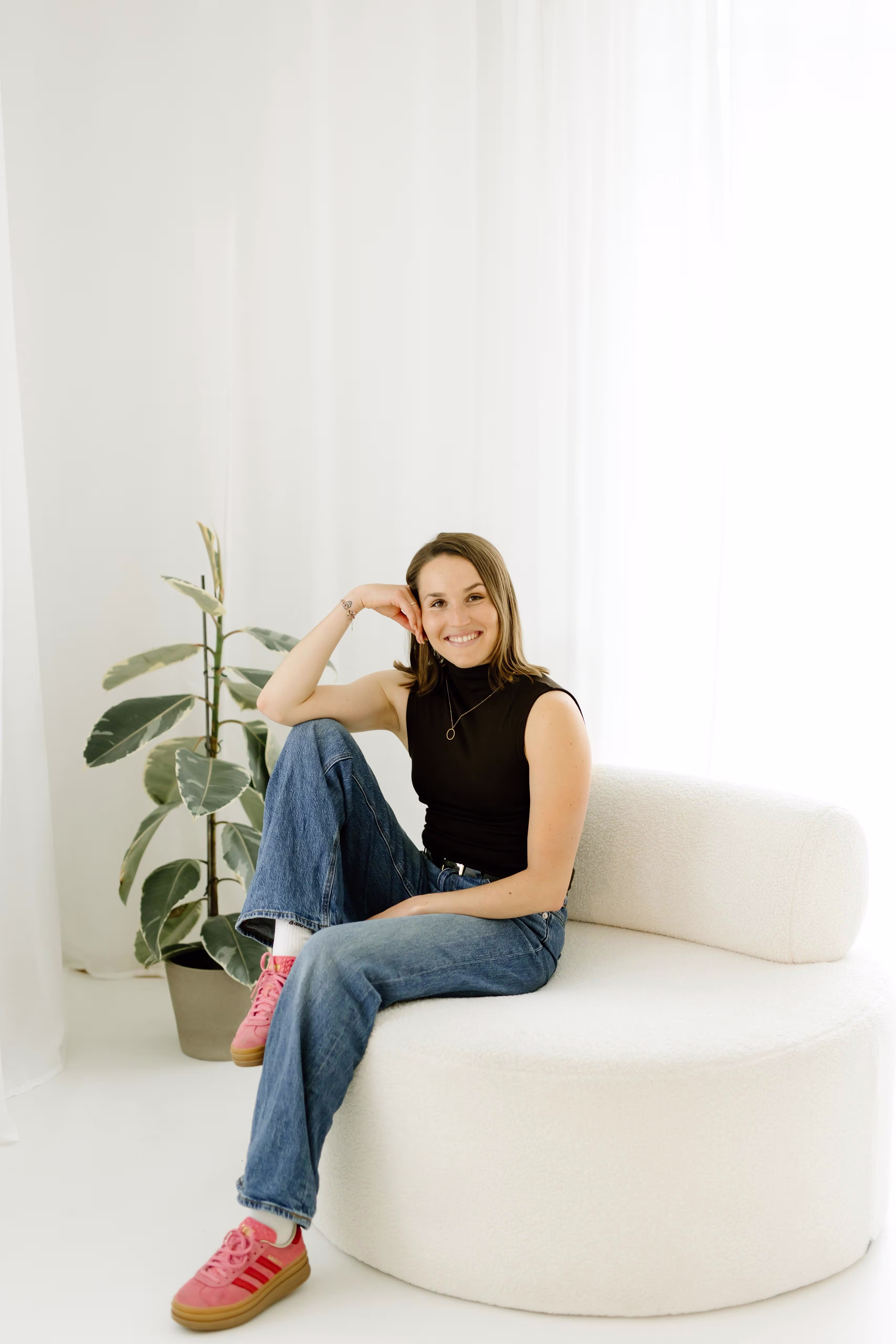 Frau sitzt lächelnd in blauer Jeans, schwarzem Oberteil und pinken Schuhen auf weißem runden Sofa.