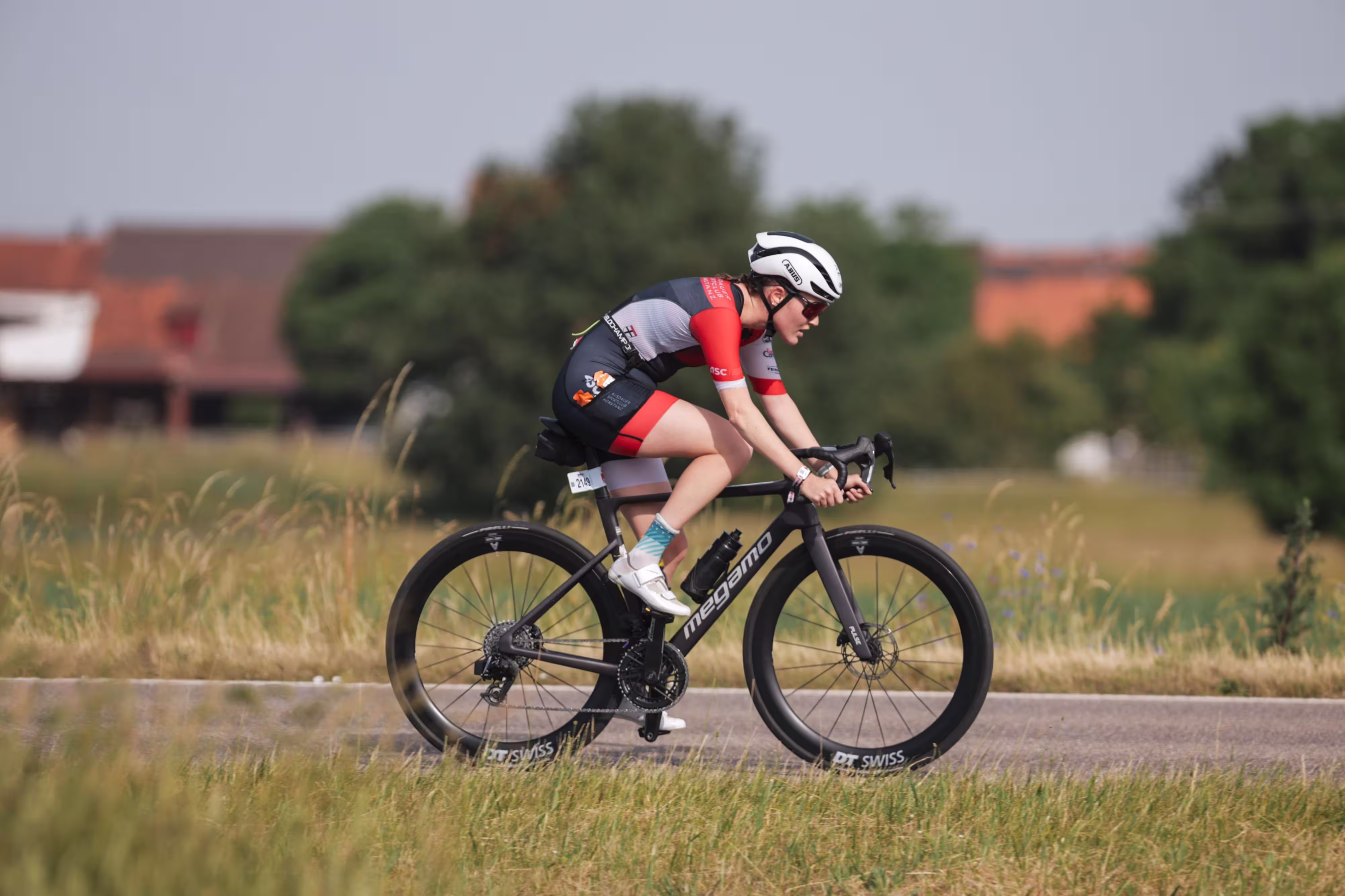 Rennradfahrerin in rotem und schwarzem Outfit fährt auf Straße durch grünes Feld.