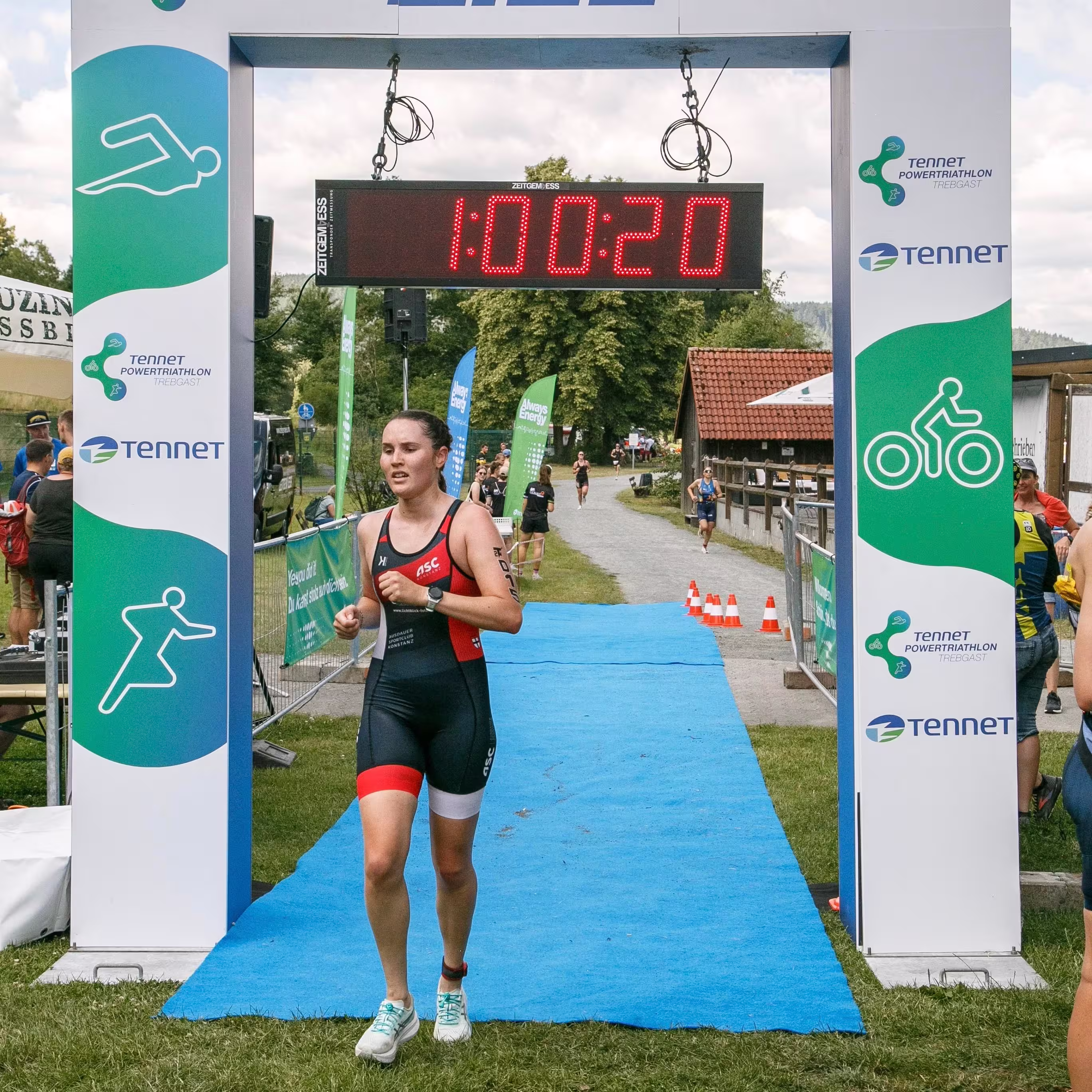 Eine Triathletin läuft über die Ziellinie bei 1:00:20 auf einer Outdoor-Strecke.