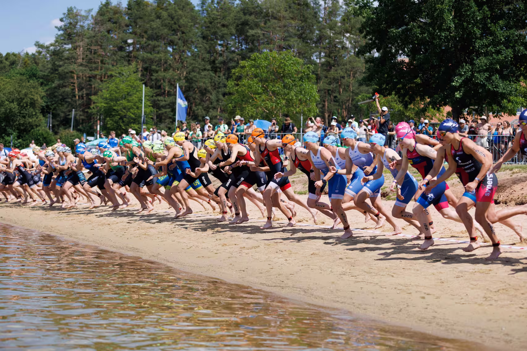 Mehrere Triathleten starten ein Schwimmrennen vom sandigen Seeufer bei sonnigem Wetter.