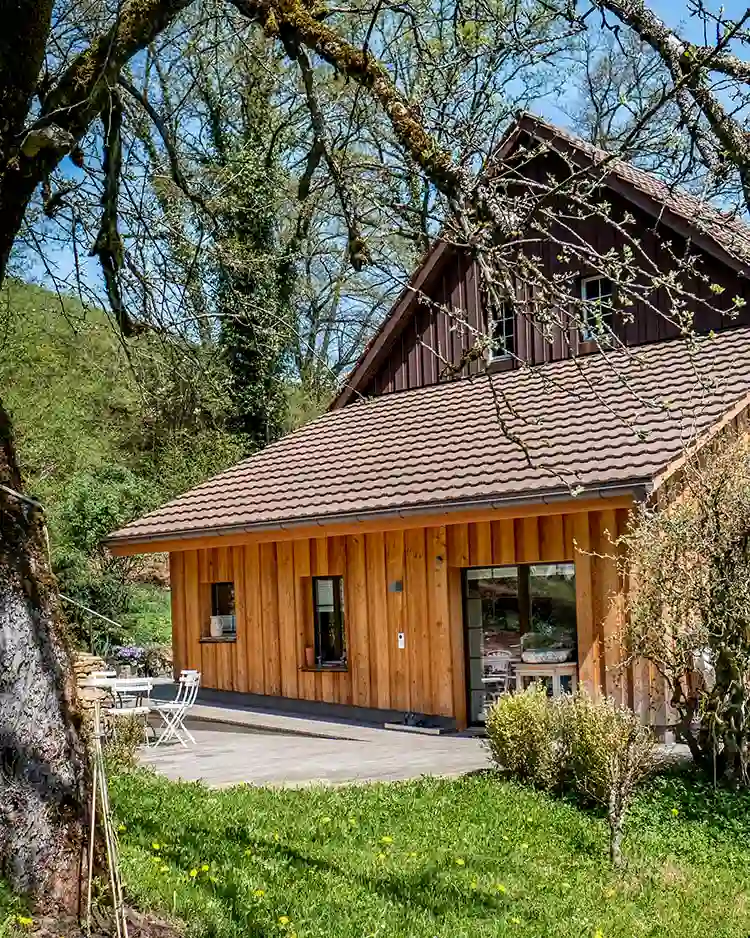 Moderner Anbau aus Holz mit Terrasse, Garten und Bäumen im Frühling unter blauem Himmel.