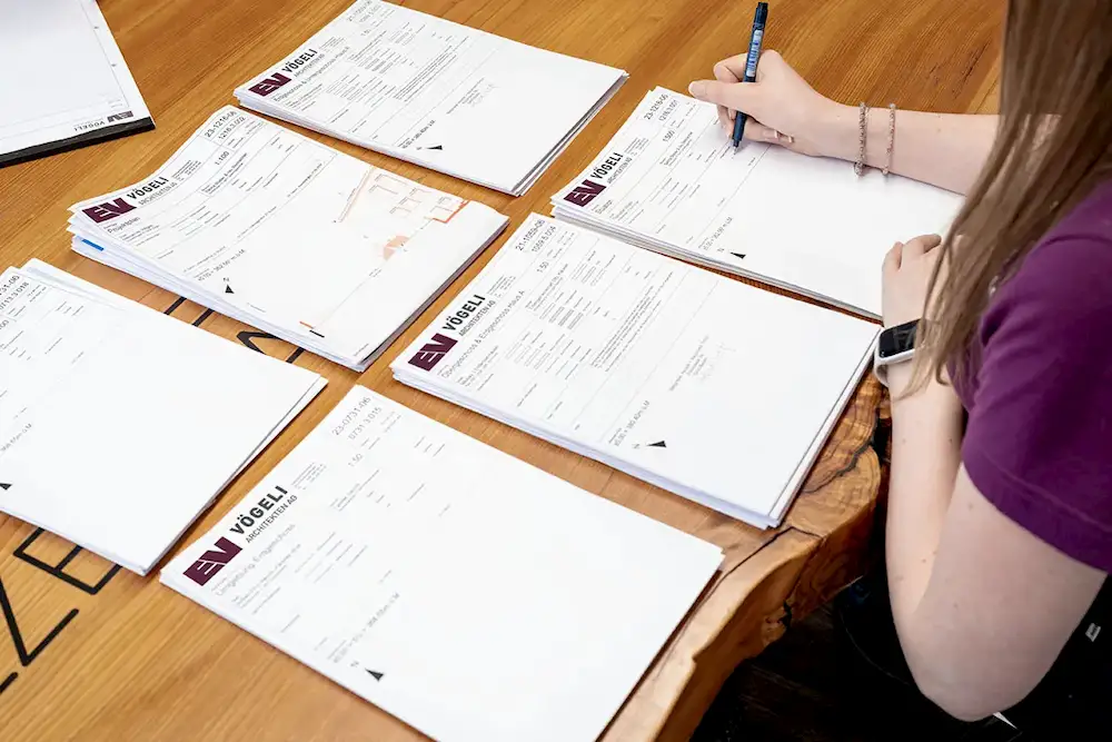 Person unterschreibt mit einem blauen Stift Pläne von Vögeli Architekten auf Holztisch.