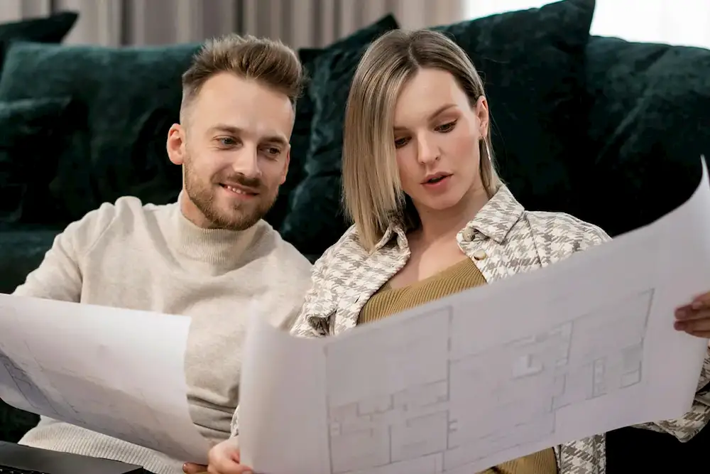 Paar sitzt auf Sofa und betrachtet gemeinsam Baupläne oder Architekturzeichnungen.