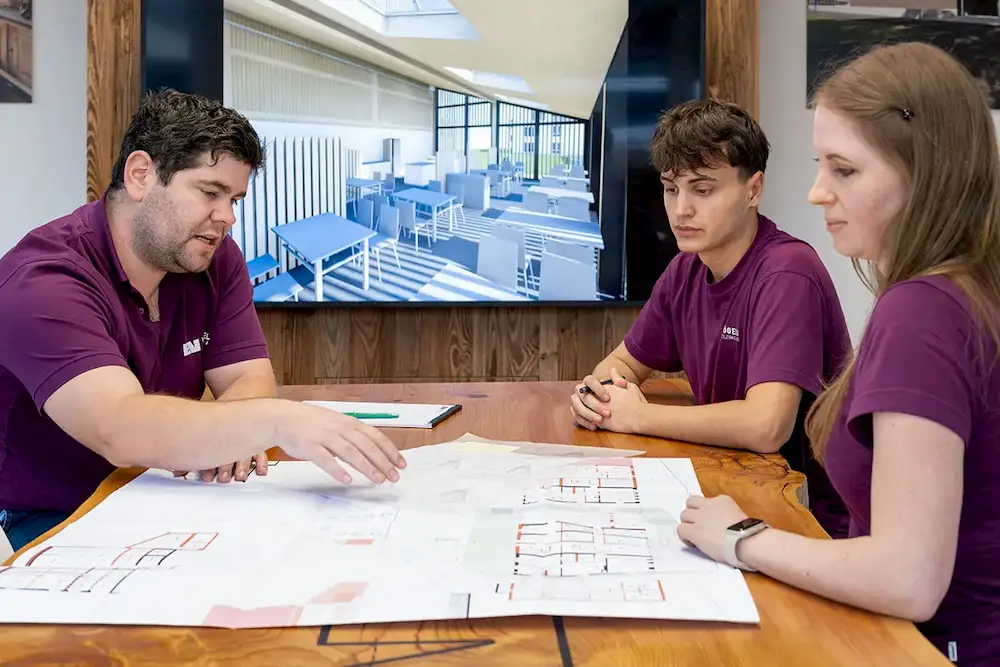 Drei Personen in violetten Shirts besprechen Architekturpläne an einem Holztisch mit 3D-Modell im Hintergrund.