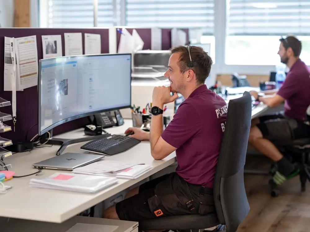 Mann in lila Shirt arbeitet an einem Schreibtisch mit gebogenem Monitor in einem Büro.