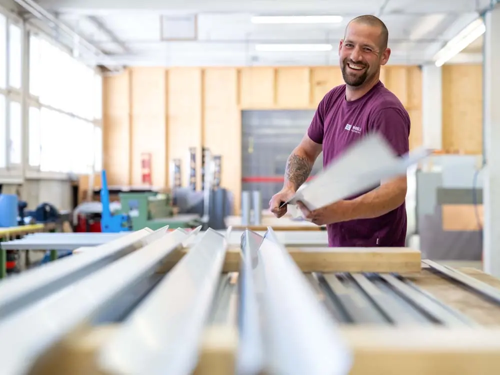 Lächelnder Arbeiter hält Metallblech in heller Werkstatt mit Maschinen und Werkzeugen.