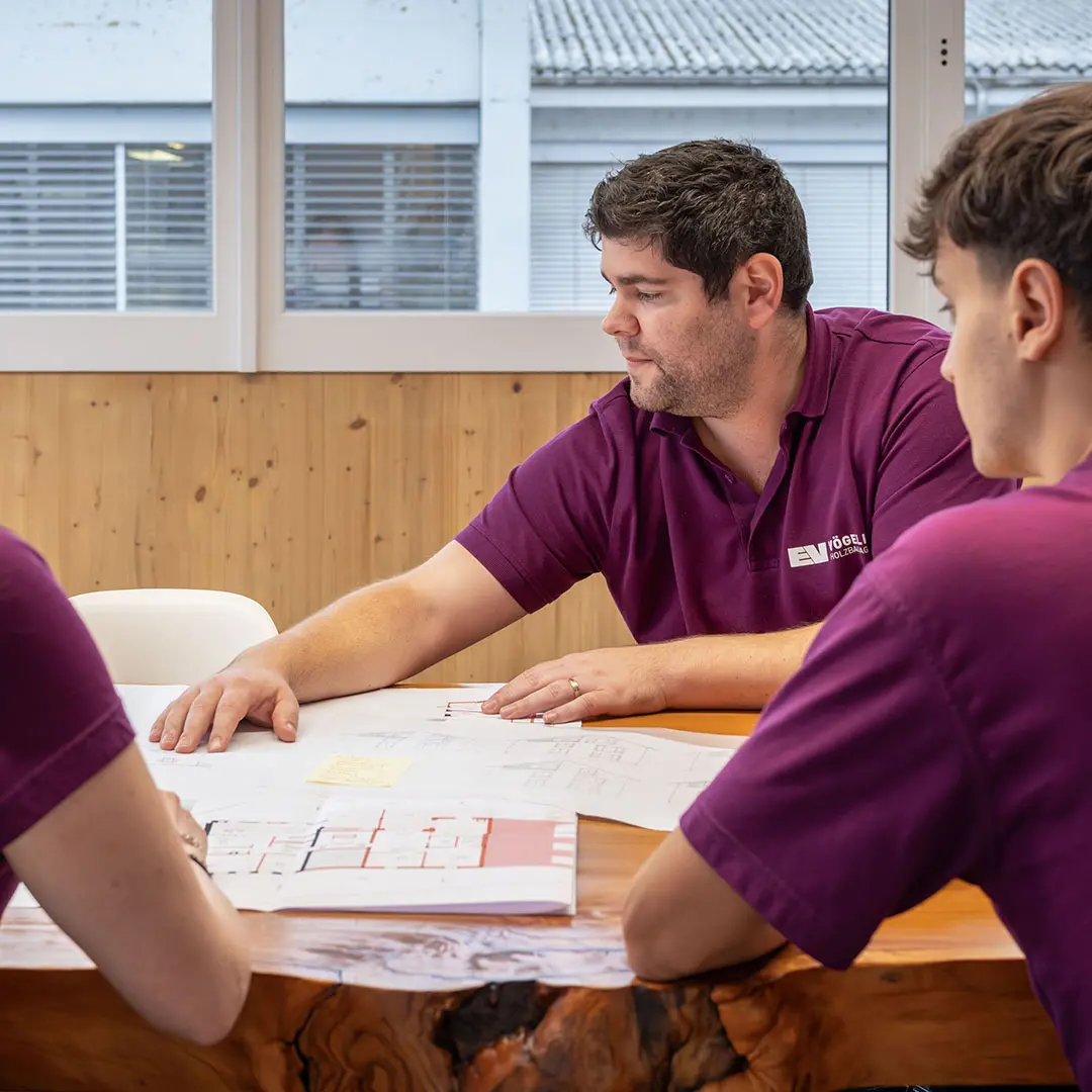 Drei Männer in violetten Poloshirts begutachten Pläne auf einem Holztisch in einem modernen Raum.