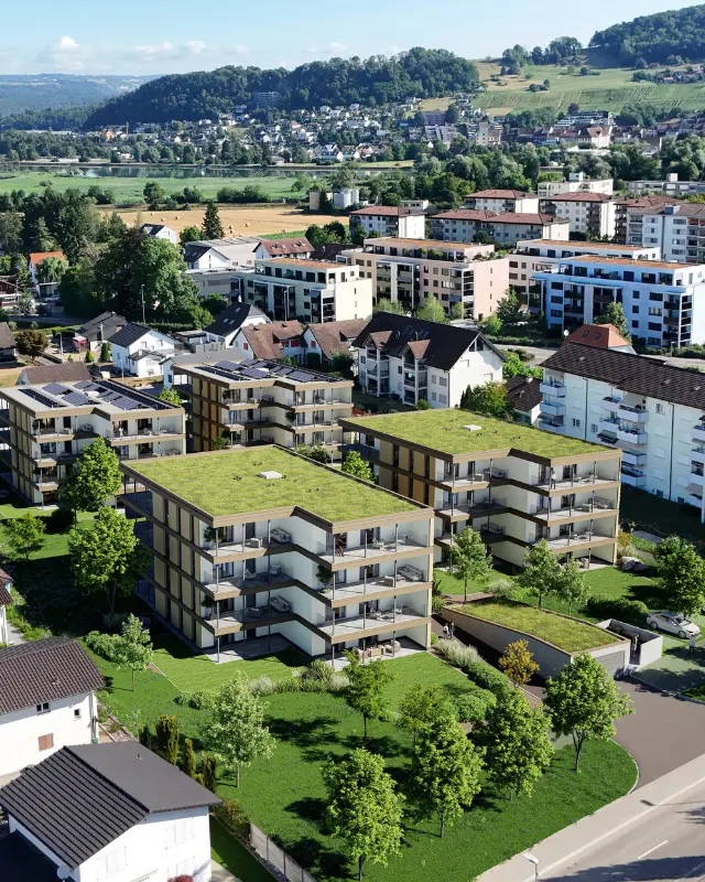 Luftaufnahme (Visualisierung) der alten Zimmerei, modernen Mehrfamilienhäusern mit begrünten Flachdächern, umgeben von gepflegtem Gras und Bäumen, im Hintergrund weitere Wohnhäuser, Felder und bewaldete Hügel.
