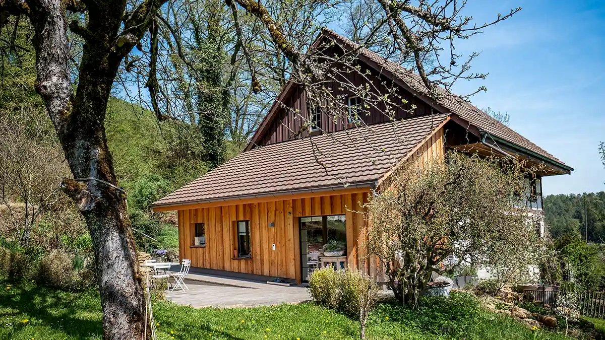 Hausanbau mit Holzfassade in ländlicher Umgebung, umgeben von Bäumen und grünem Rasen, bei klarem Himmel.