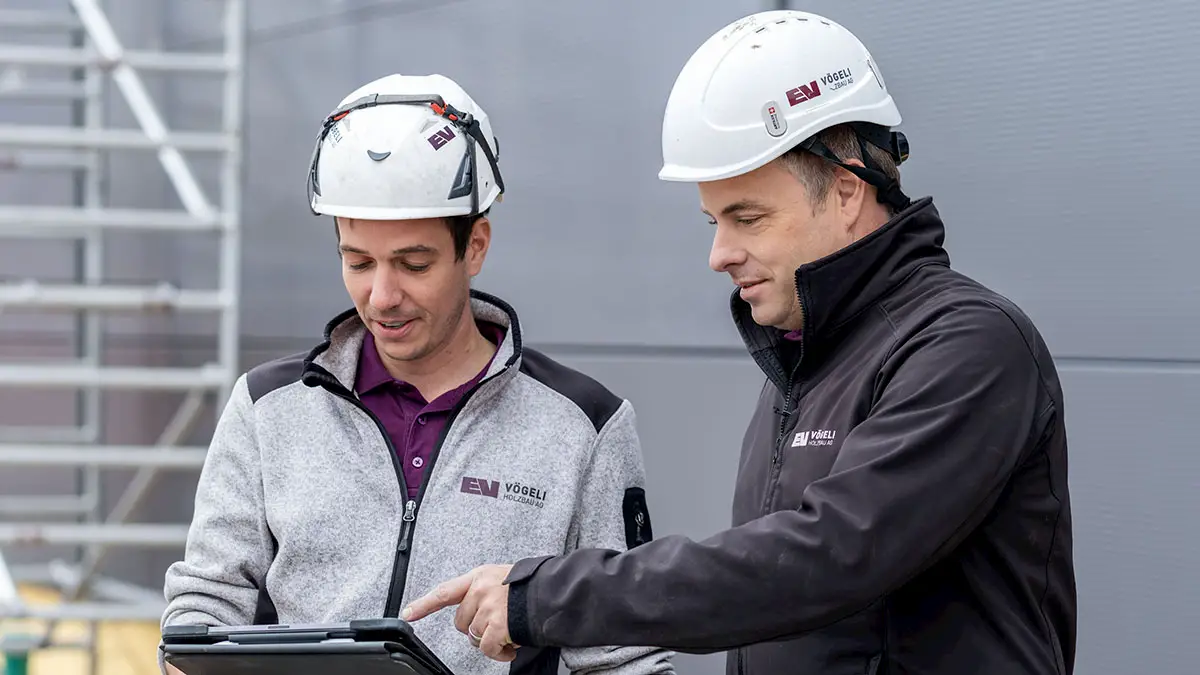 Bauleiter der Vögeli Architekten auf einer Baustelle tragen weisse Schutzhelme und VöGELI Architekten Arbeitskleidung, einer hält ein Tablet, das andere zeigt darauf.