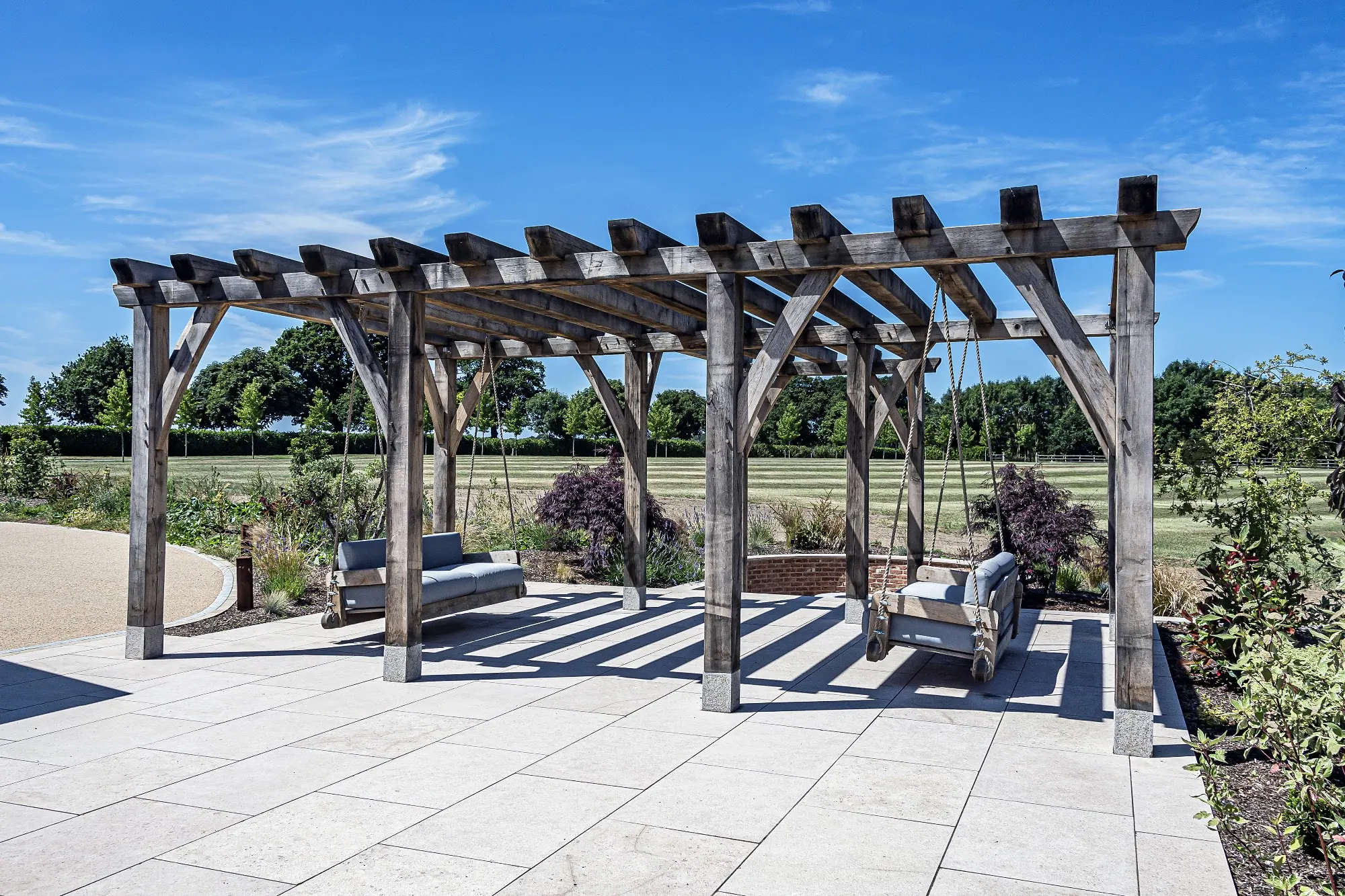 The oak pergola at Ullenhall, Warwickshire, with two hanging oak sofas under it on a paved terrace