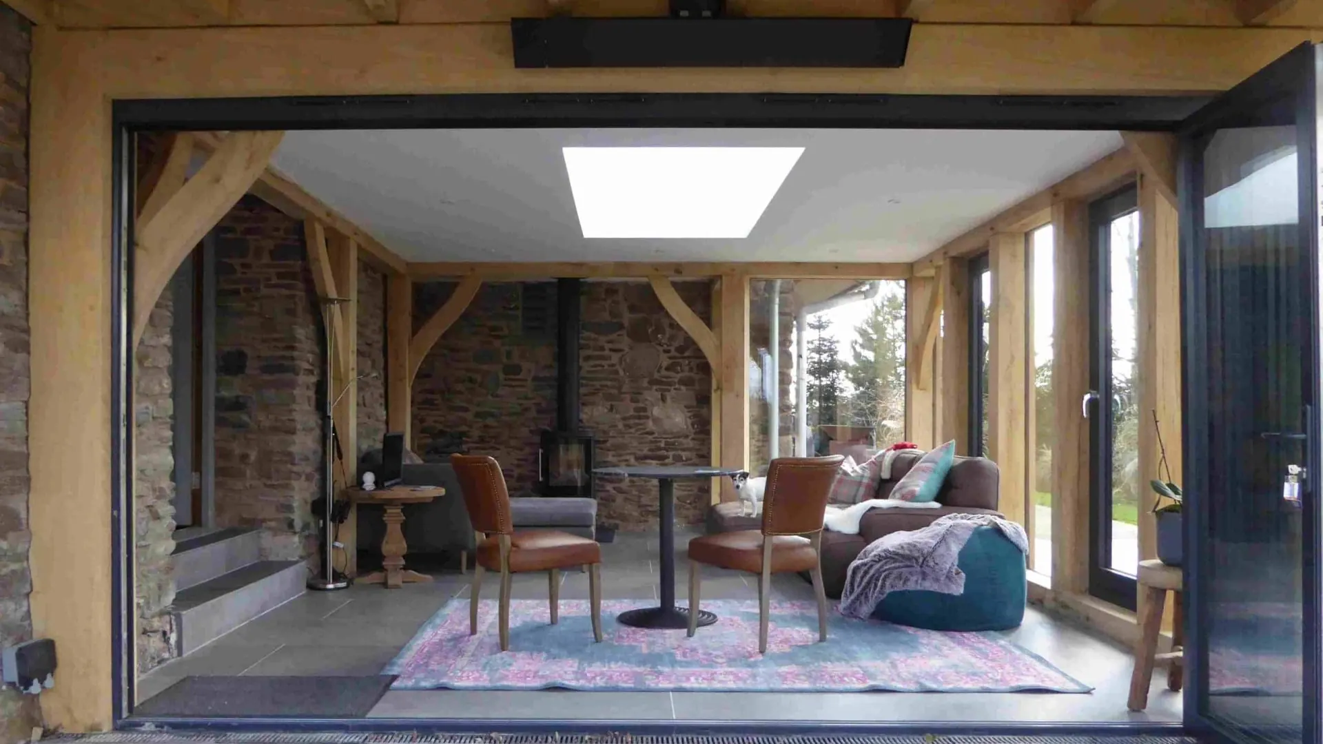 Looking in through open bifold doors at the flat-roofed oak frame living space at Llanvetherine, with its roof light and the stone barn wall inside