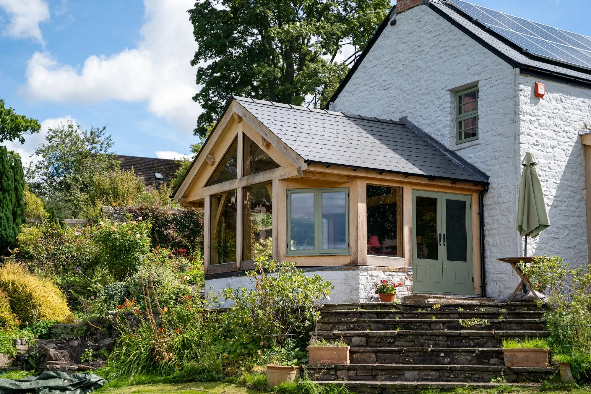 The oak frame extension at Ty Fry, Crickhowell: a glazed gable on the end of a white-painted stone house, above steps down to the garden.