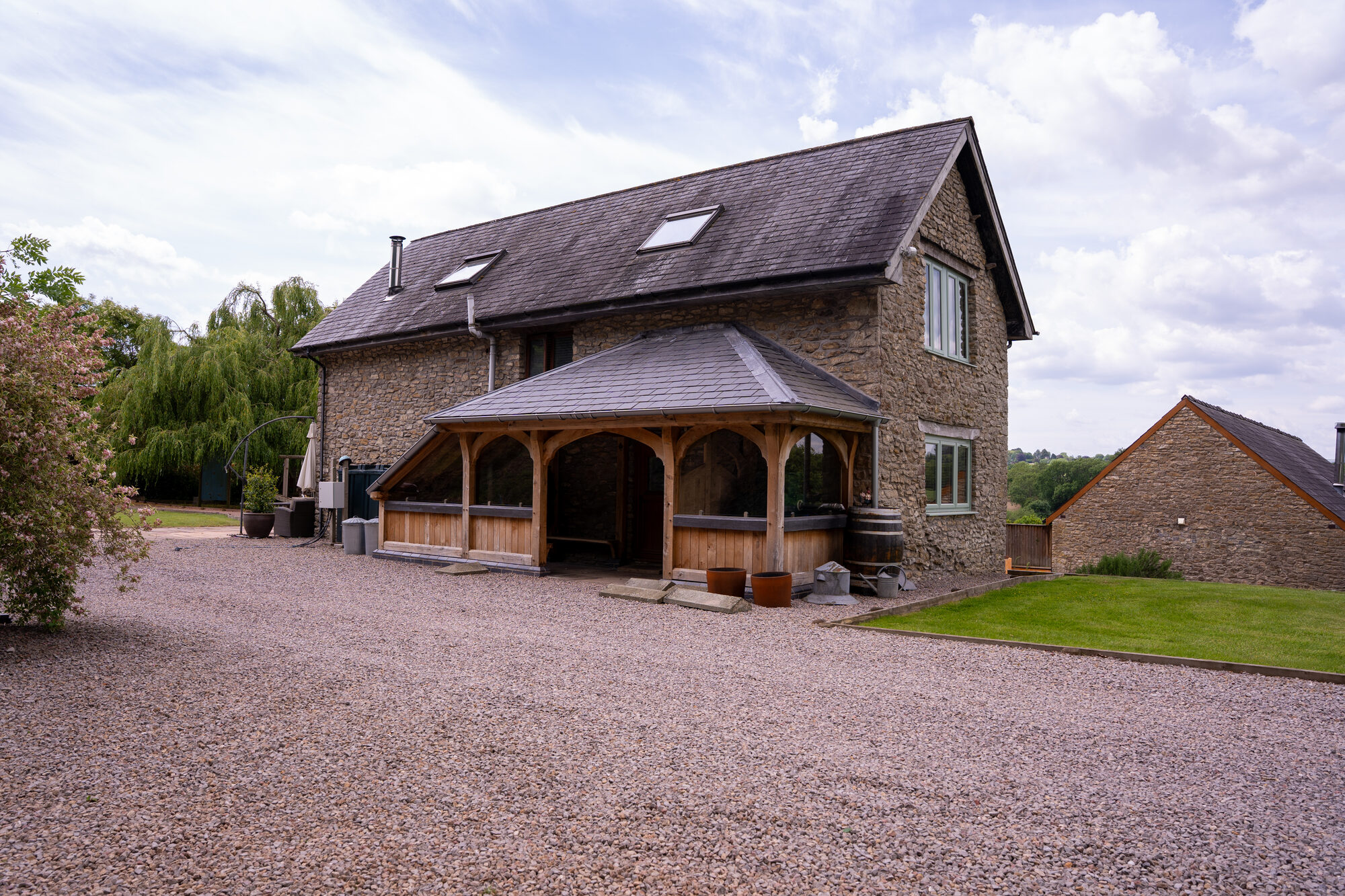 Open oak frame porch with a hipped slate roof against a stone barn near Usk