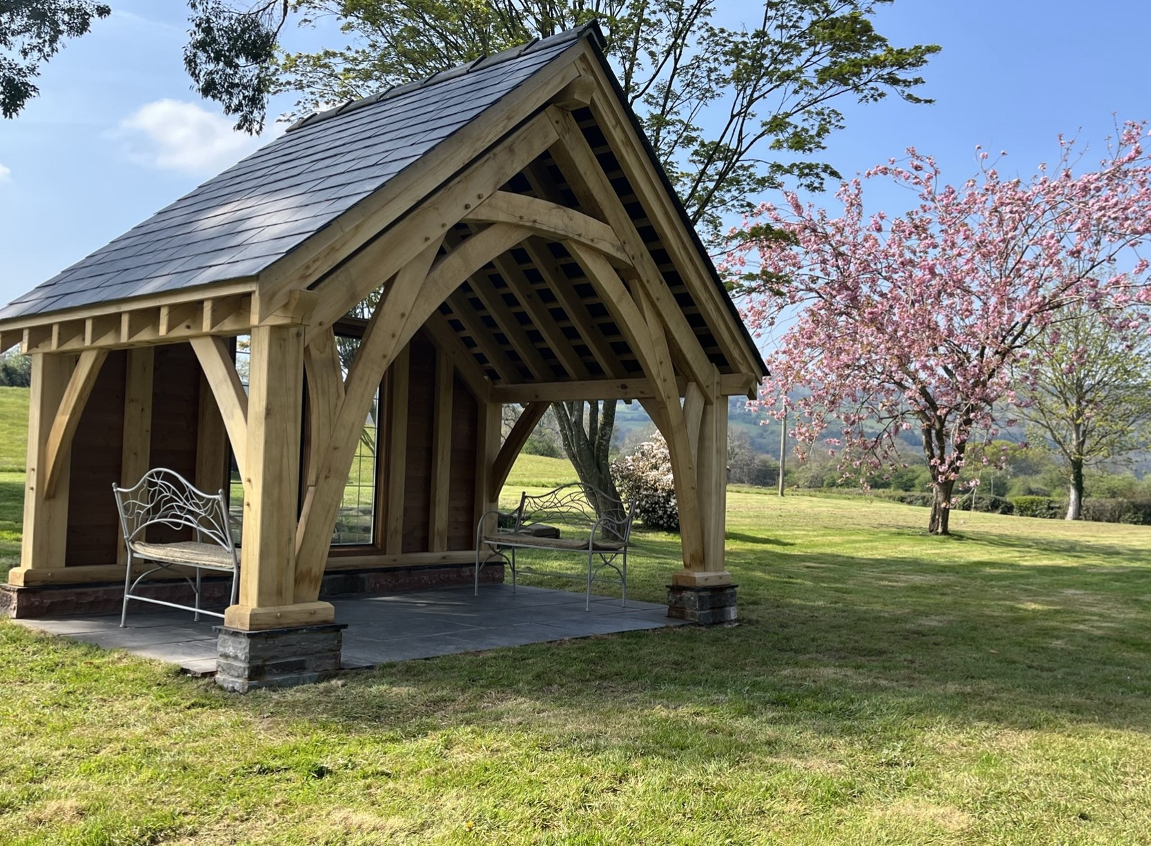 Oak wedding gazebo at Duffryn Mawr, Gilwern, with a slate roof and exposed oak battens, standing on the lawn beside a cherry tree in blossom
