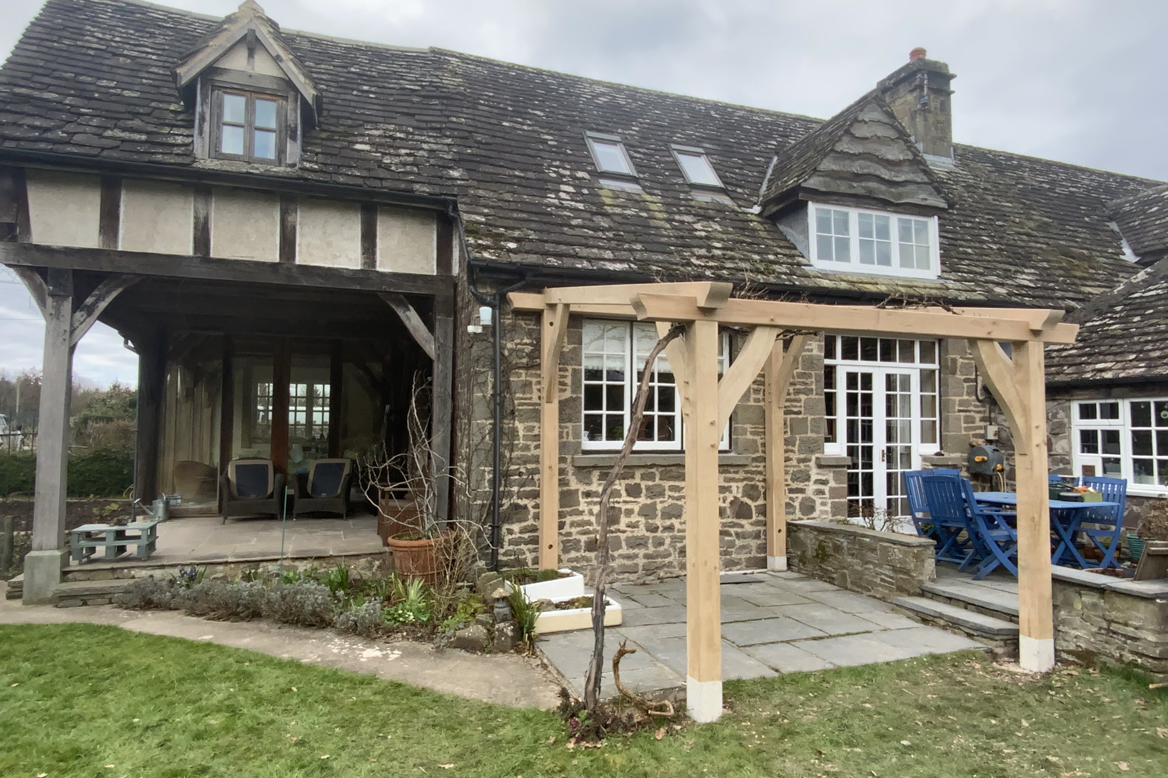 Oak pergola with curved braces on a stone terrace behind a cottage