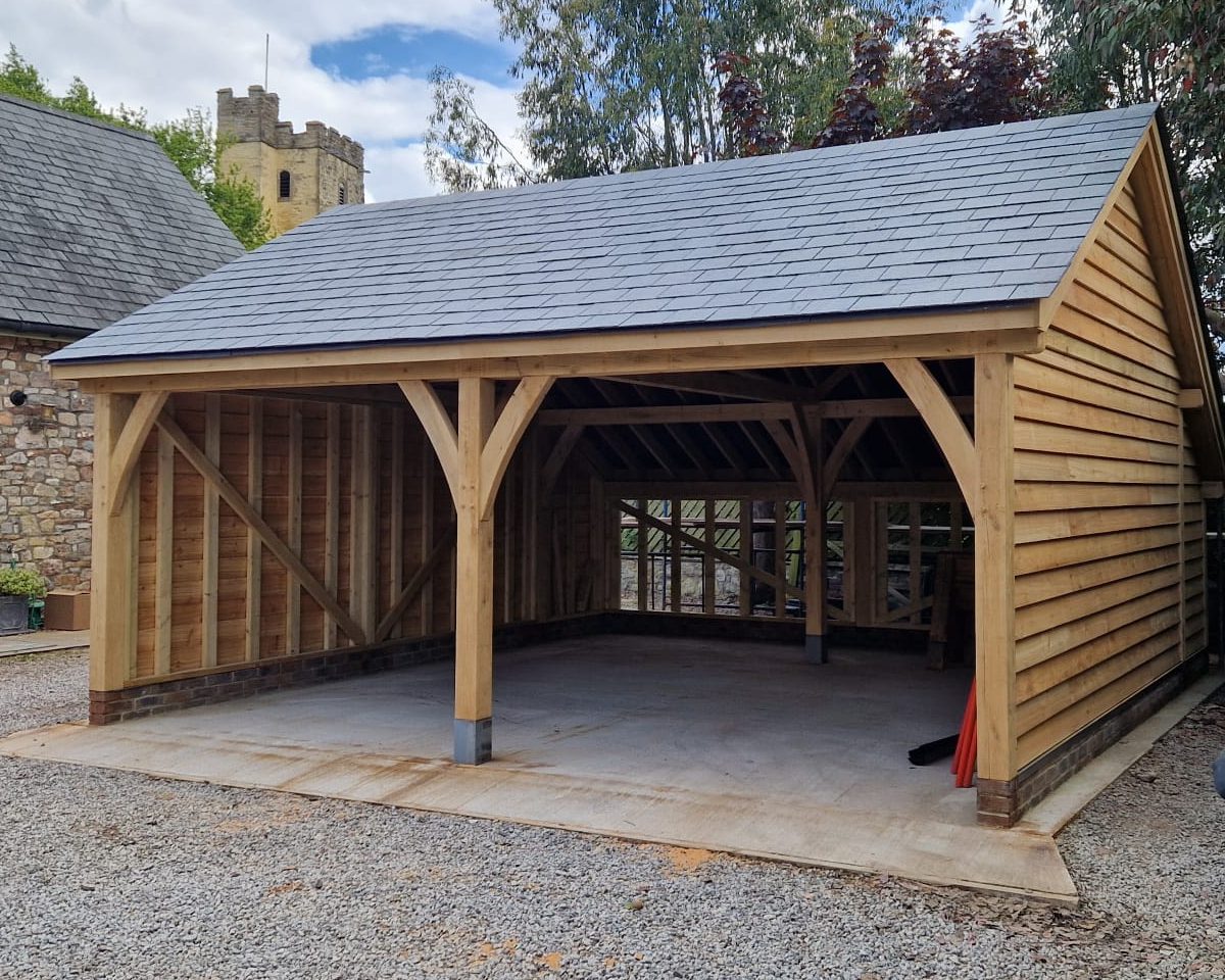 A two-bay open-fronted oak frame garage with featheredge-clad sides and a slate roof, on a gravel drive.