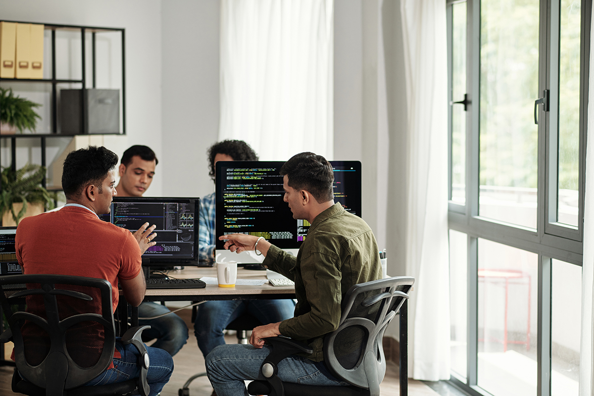 Freelance developers collaborating at a desk with multiple monitors displaying code in a bright office