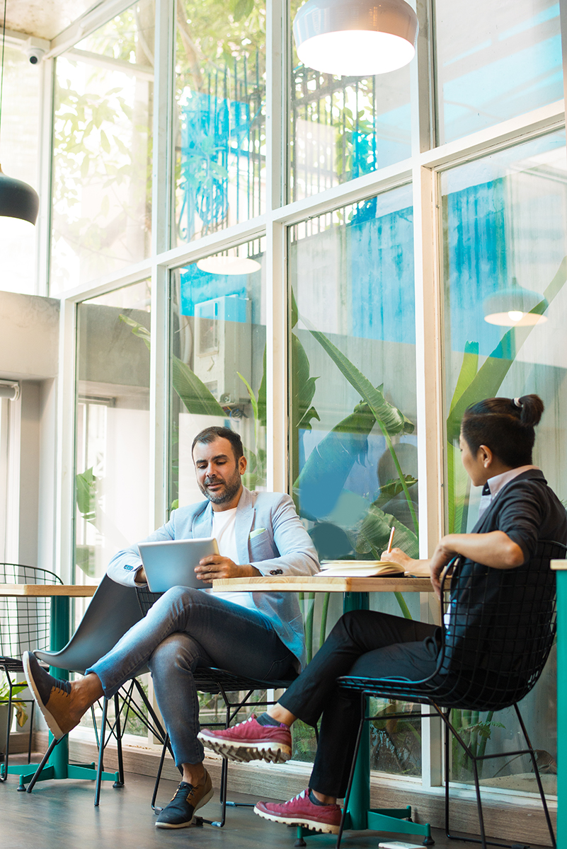 Two professionals collaborating at a table in a bright, plant-filled workspace, one holding a tablet and the other taking notes in a notebook