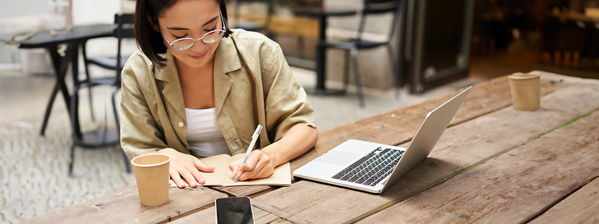 Freelancer writing notes in a notebook at a café table with a laptop, smartphone, and coffee nearby