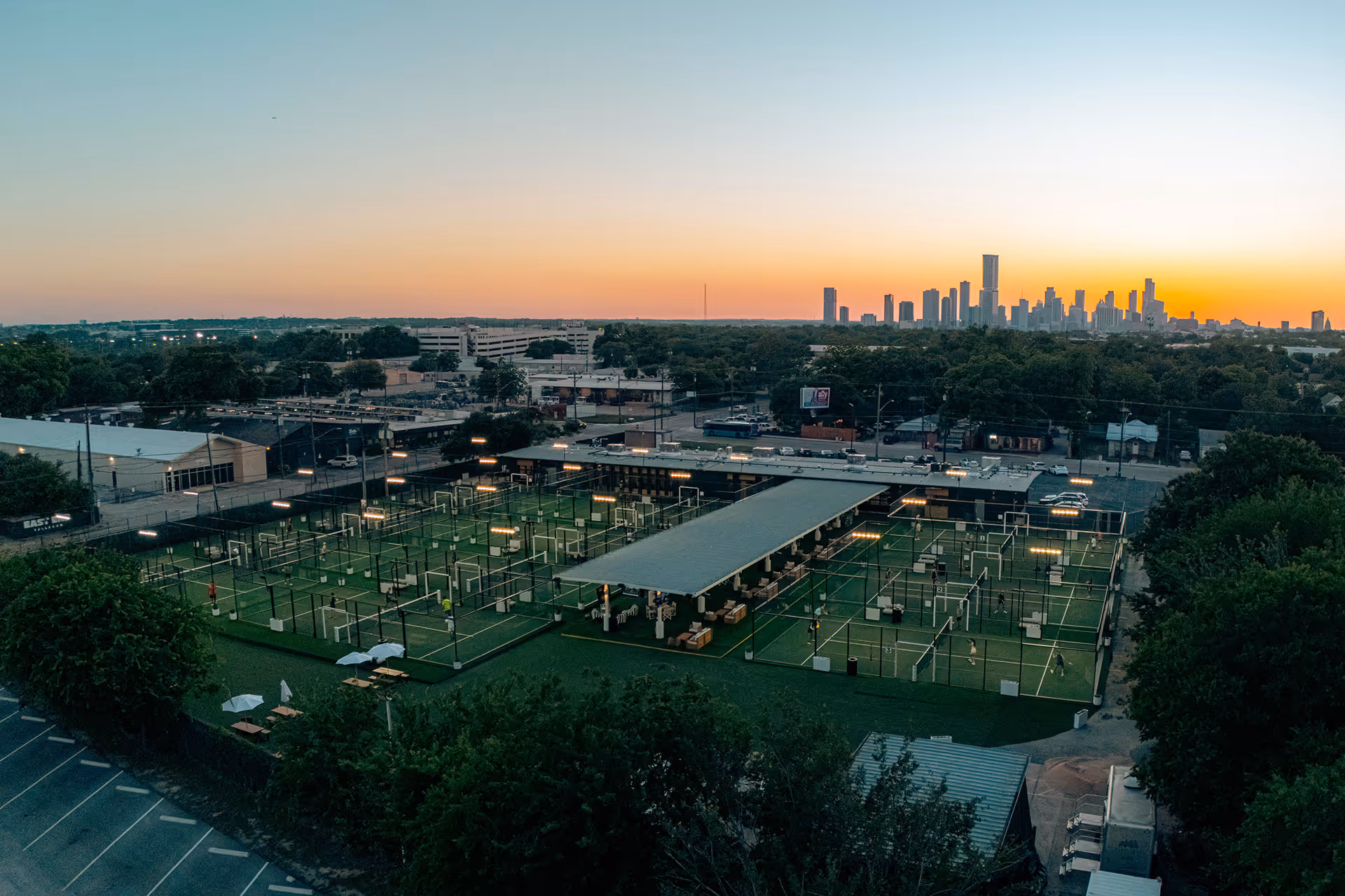 Aerial view of multiple illuminated padel courts with players at sunset and the Austin, Texas, city skyline in the background.